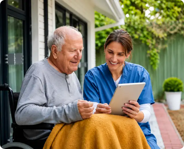 A smiling female nurse in blue scrubs showing a digital tablet screen to an elderly man wrapped in a blanket in a wheelchair outdoors.
