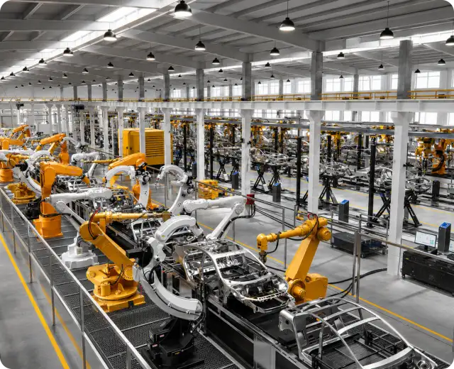 Wide overhead view of a modern automobile factory floor featuring multiple yellow and white robotic arms assembling metal car chassis along a production line.