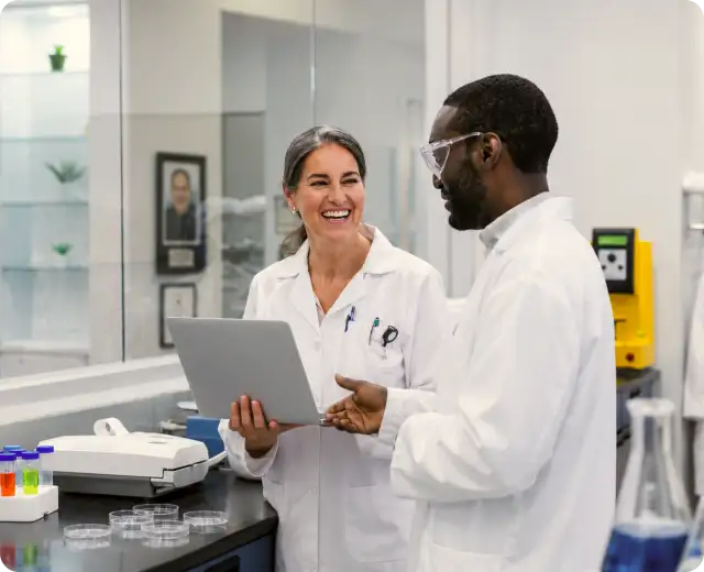 A female and a male scientist in lab coats smiling and discussing data on a laptop inside a laboratory equipped with petri dishes and test tubes.