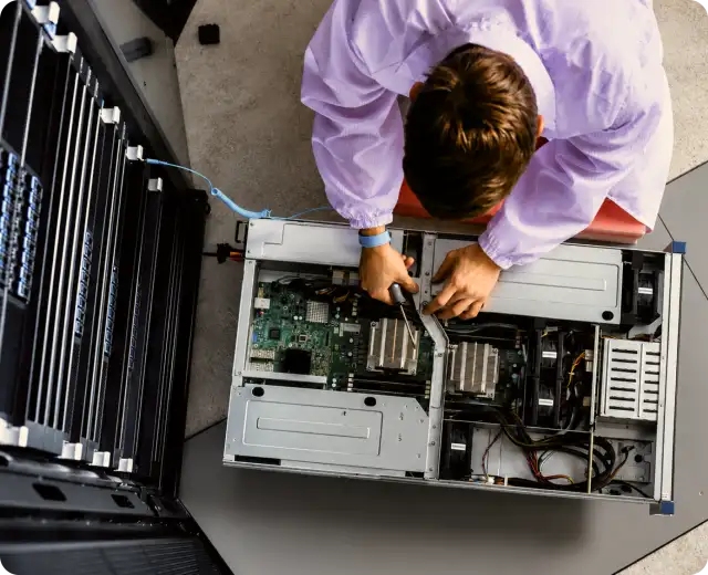 Overhead view of an IT technician using a screwdriver to repair or assemble components inside an open server chassis next to a server rack.