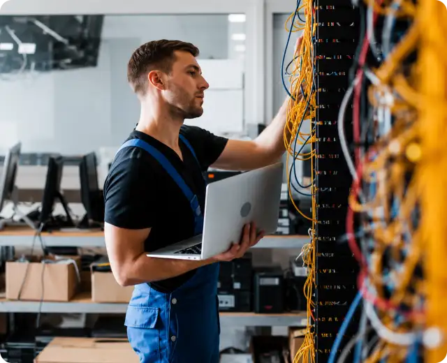 An IT technician in blue overalls holding an open laptop while inspecting complex network and server wiring in a data center or server room.