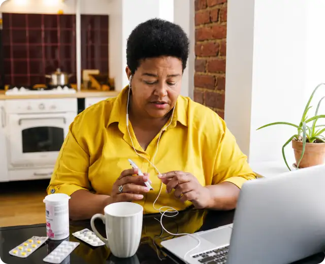 A woman sitting at a kitchen table wearing earphones, looking at an open laptop with prescription pill bottles and blister packs nearby.