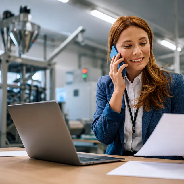 Woman on a call reviewing paperwork at her laptop on a manufacturing floor.